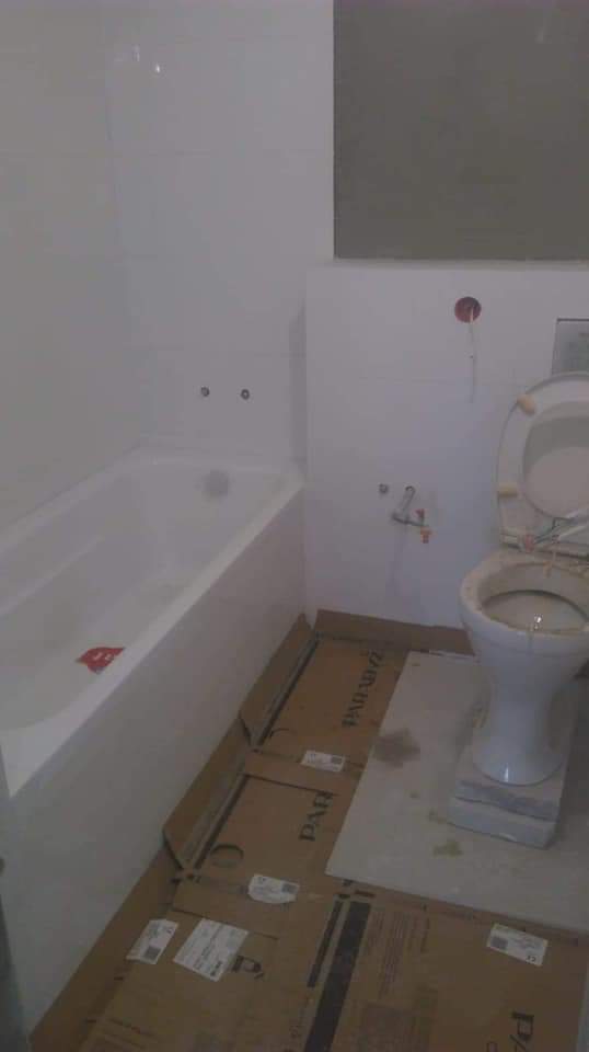 Łazienka w trakcie remontu: biała wanna zabudowana płytkami, toaleta z otwartą klapą, podłoga zabezpieczona kartonem, widoczne niedokończone przyłącza wodne i elektryczne.