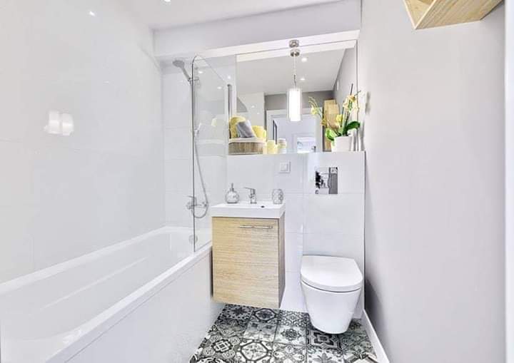 Nowoczesna, jasna łazienka z białą wanną za szklanym parawanem, szafką pod umywalką z drewnianym frontem, toaletą typu Geberit i wzorzystą, czarno-białą podłogą.