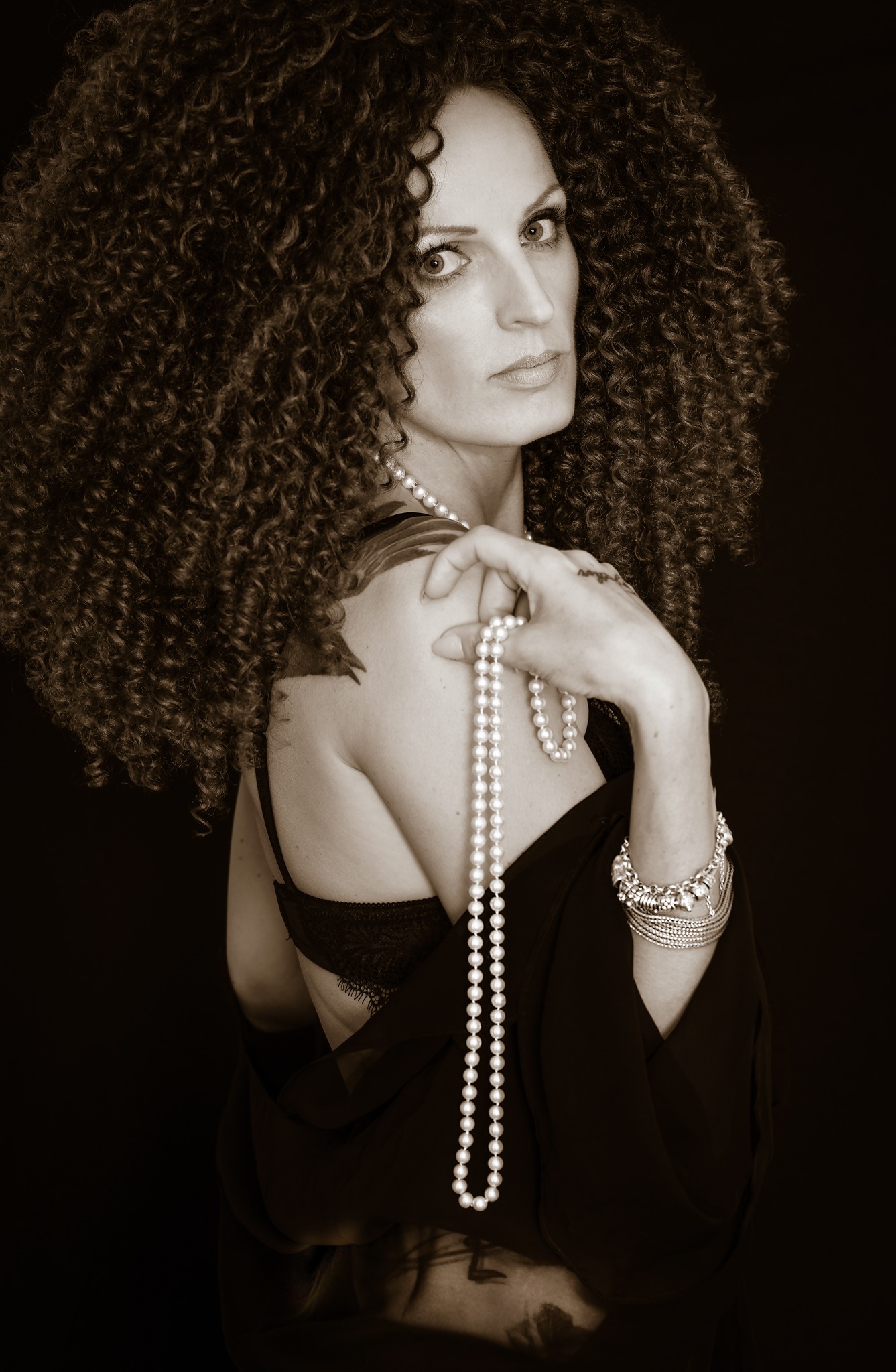 Portret kobiety z burzą loków, perłami i bransoletkami, pozującej na ciemnym tle, ujęcie od ramienia w górę.