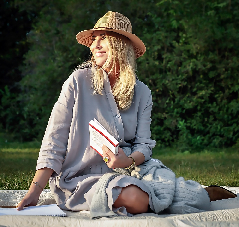 Uśmiechnięta blondynka w kapeluszu, siedząca na kocu na trawie, trzymająca książkę z czerwoną okładką, w tle zielone drzewa.