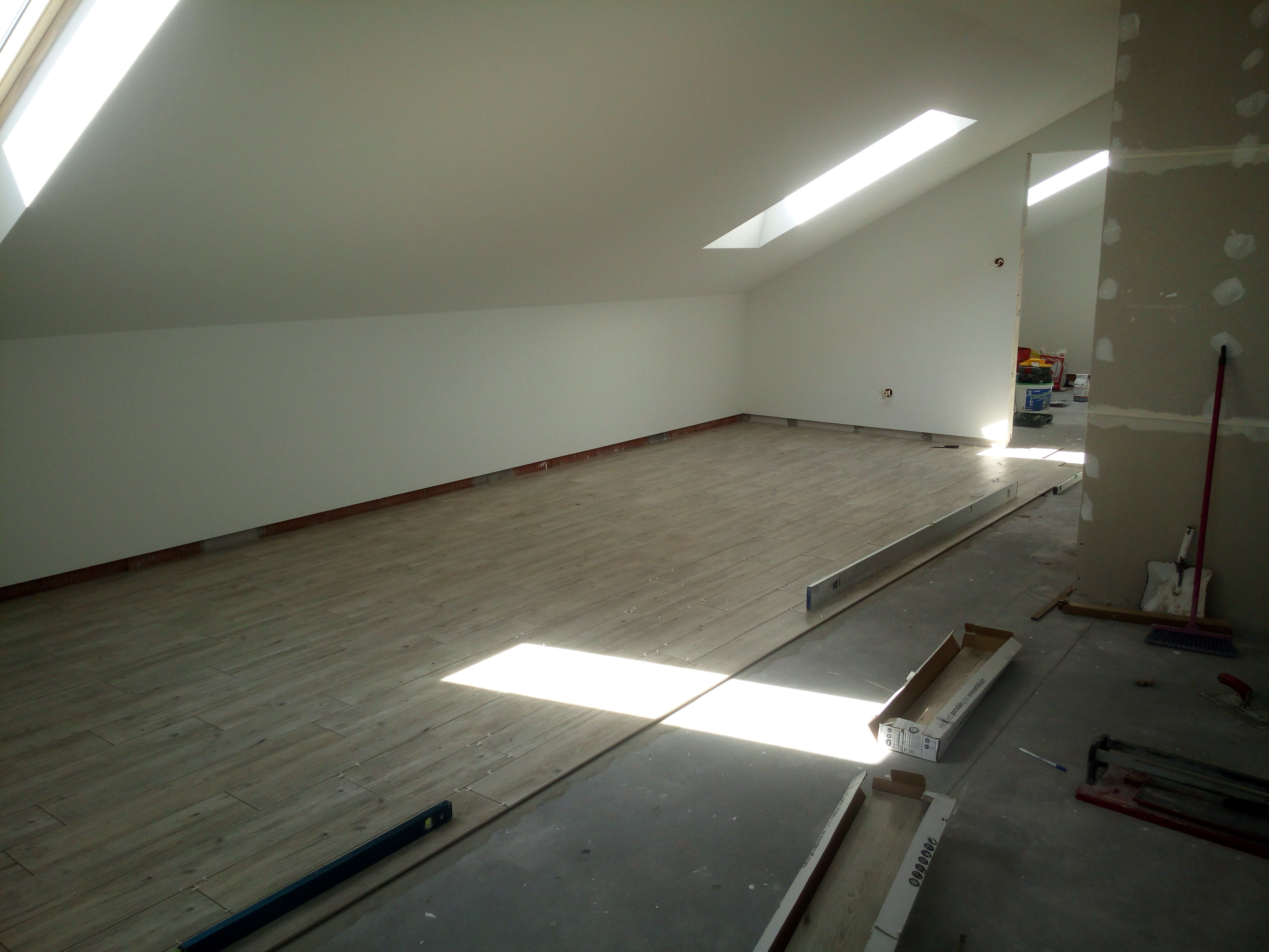 Poddasze w trakcie układania podłogi z płytek imitujących drewno, widoczne narzędzia glazurnicze, poziomica i kartony po płytkach.