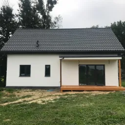 Nowoczesny, parterowy dom z białymi ścianami, grafitowym dachem i drewnianym tarasem, widok z zewnątrz w pochmurny dzień.