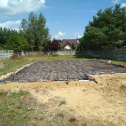 Przygotowanie zbrojenia płyty fundamentowej na czarnej folii, obłożonej cegłami i rurami kanalizacyjnymi, widok z poziomu gruntu na tle zieleni i betonowego ogrodzenia.