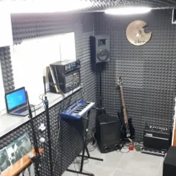 Wnętrze studia nagrań z panelami akustycznymi, gitarą basową, keyboardem, mikrofonami, wzmacniaczem VOX i talerzem perkusyjnym Zildjian na ścianie.
