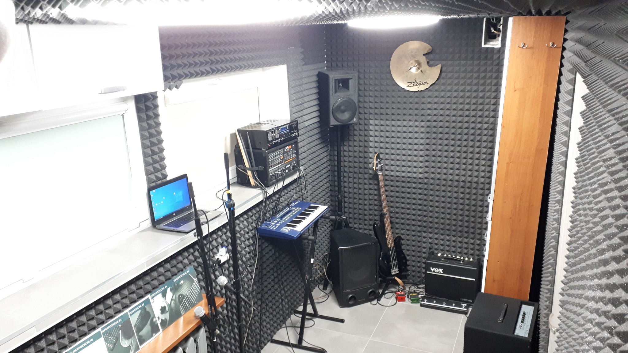 Wnętrze studia nagrań z panelami akustycznymi, gitarą basową, keyboardem, mikrofonami, wzmacniaczem VOX i talerzem perkusyjnym Zildjian na ścianie.