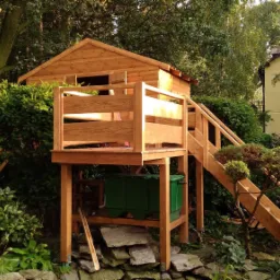 Drewniany domek na drzewie z tarasem i schodami, umieszczony wśród zieleni, z widocznym zbiornikiem na wodę pod konstrukcją.