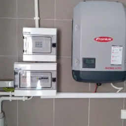 Szara ściana z ceramicznych płytek, na której zamontowano dwa białe rozdzielnice elektryczne, szary inwerter Fronius Symo z wyświetlaczem oraz częściowo widoczny, biały zasobnik ciepłej wody Buderus.