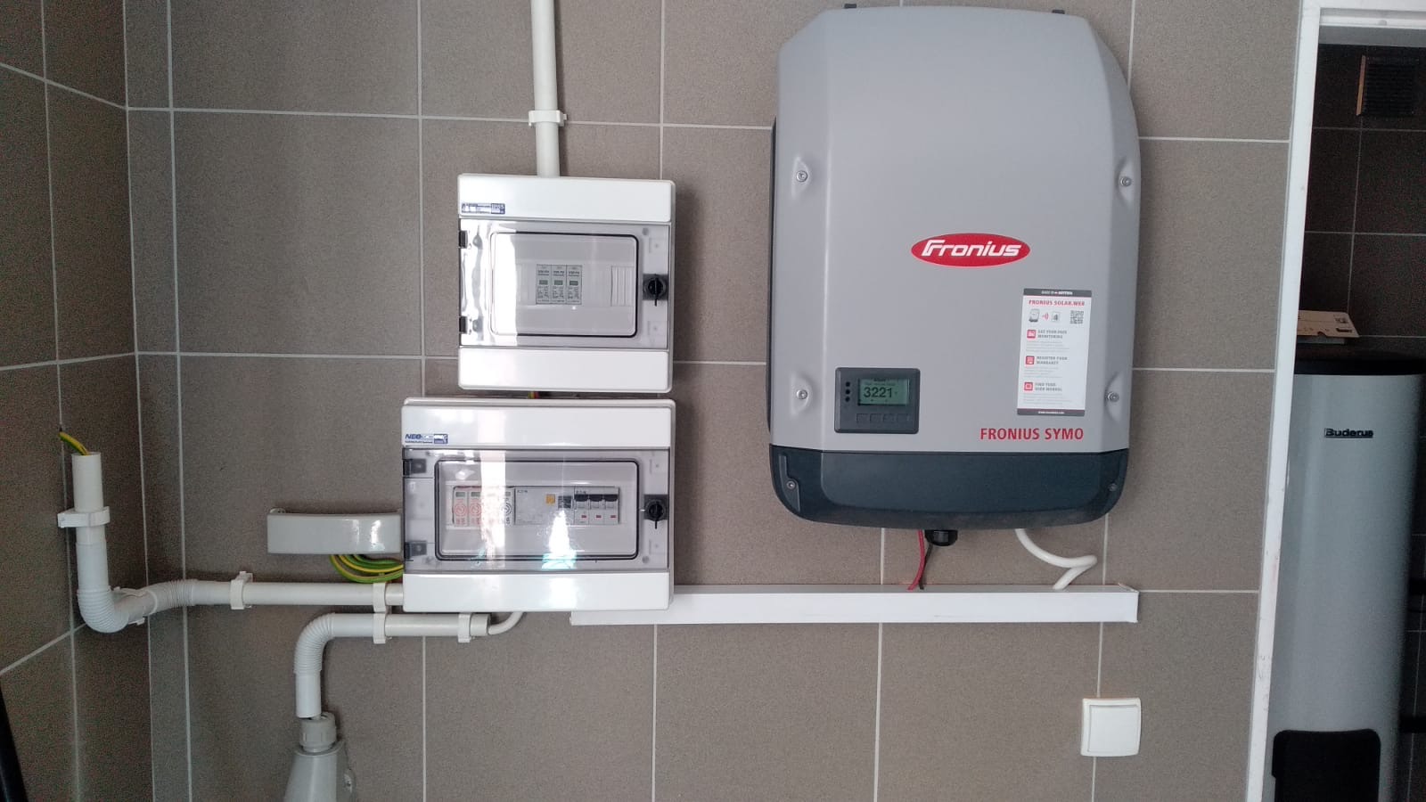 Szara ściana z ceramicznych płytek, na której zamontowano dwa białe rozdzielnice elektryczne, szary inwerter Fronius Symo z wyświetlaczem oraz częściowo widoczny, biały zasobnik ciepłej wody Buderus.
