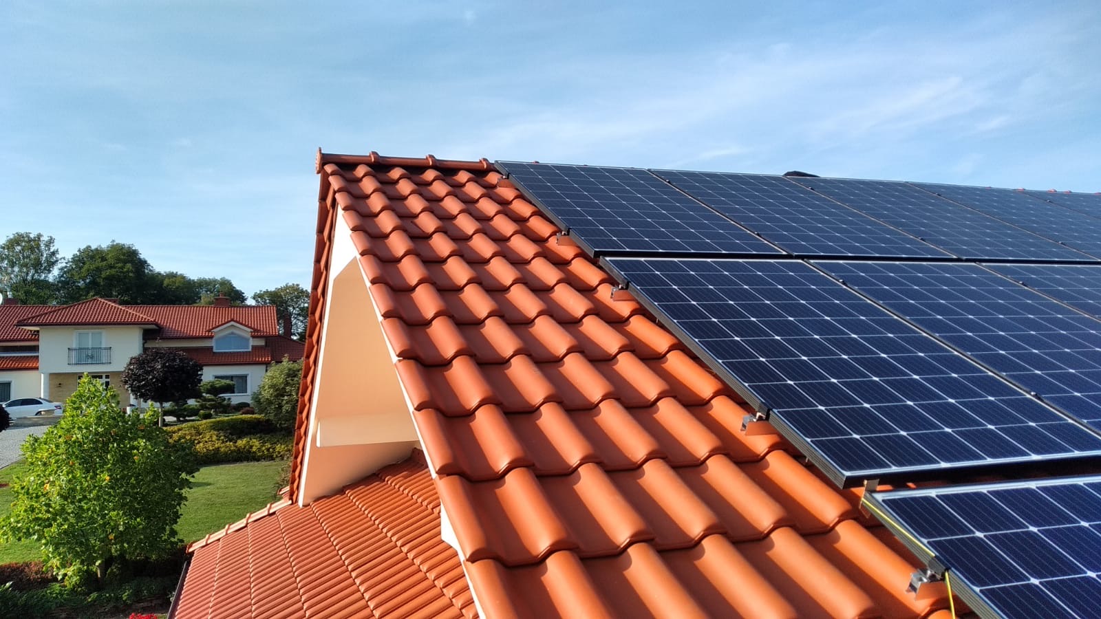 Czerwona dachówka ceramiczna z zainstalowanymi panelami słonecznymi, widok z góry na dom z ogrodem w tle pod błękitnym niebem.