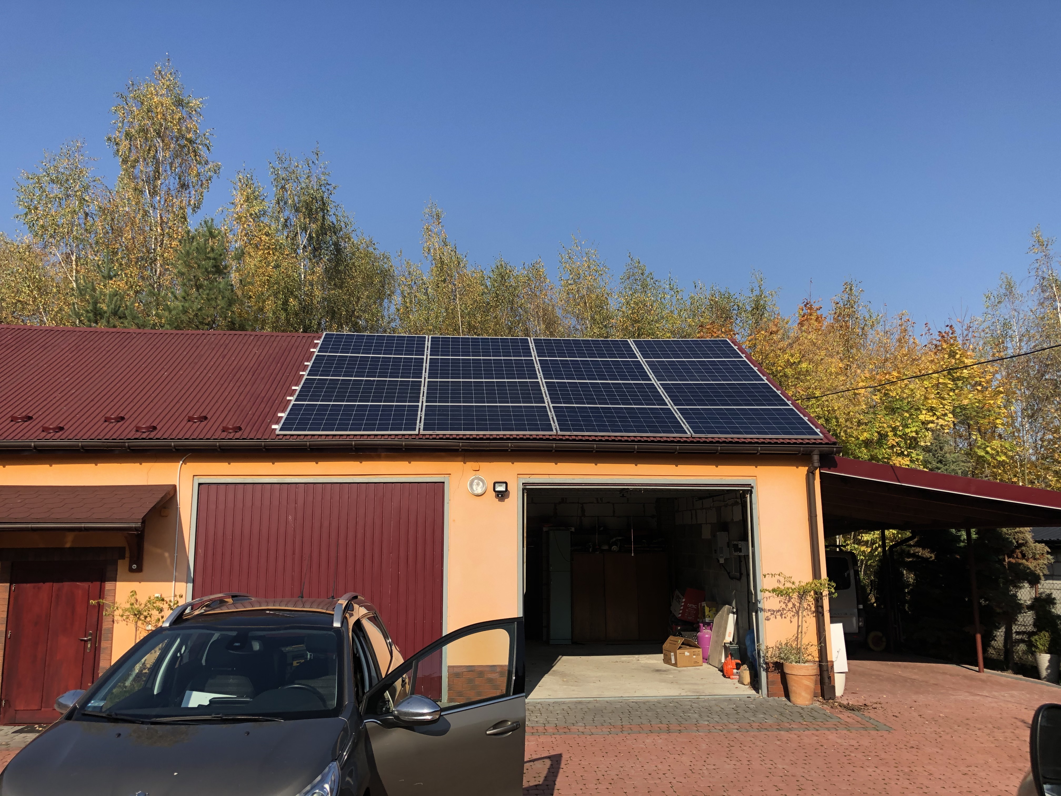 Panele słoneczne na dachu pomarańczowego garażu z otwartymi drzwiami i zaparkowanym samochodem z otwartymi drzwiami na tle jesiennych drzew i błękitnego nieba.