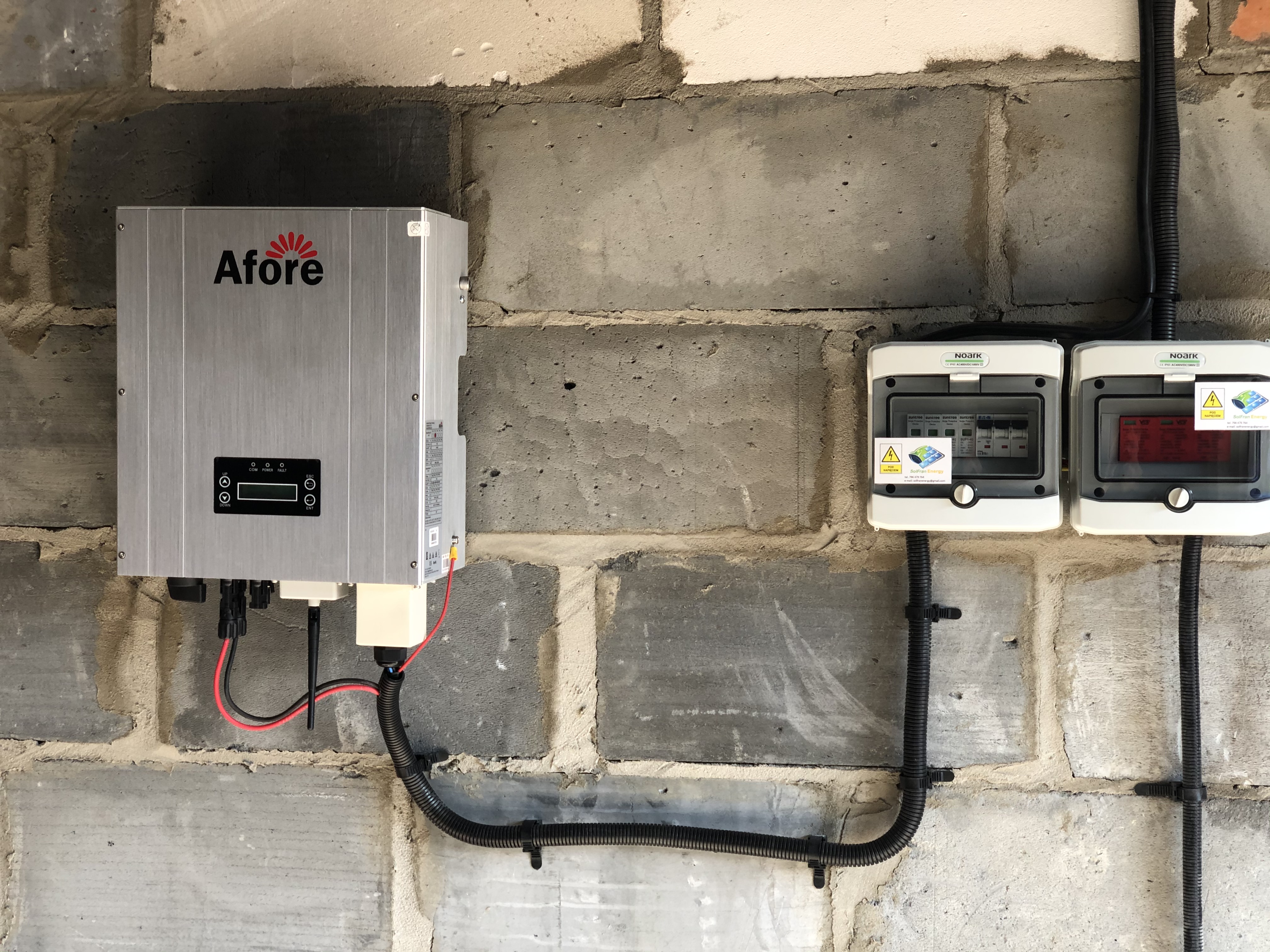 Szary inwerter firmy Afore zamontowany na surowej ścianie z bloczków, obok dwie białe skrzynki elektryczne NOARK z widocznymi bezpiecznikami i naklejkami ostrzegawczymi, przewody w peszlach...