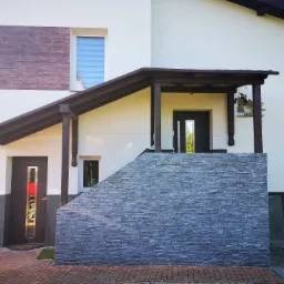 Elewacja domu z kamienną okładziną schodów wejściowych i drewnianym panelem dekoracyjnym, widoczne okna z roletami i zadaszenie nad wejściem.