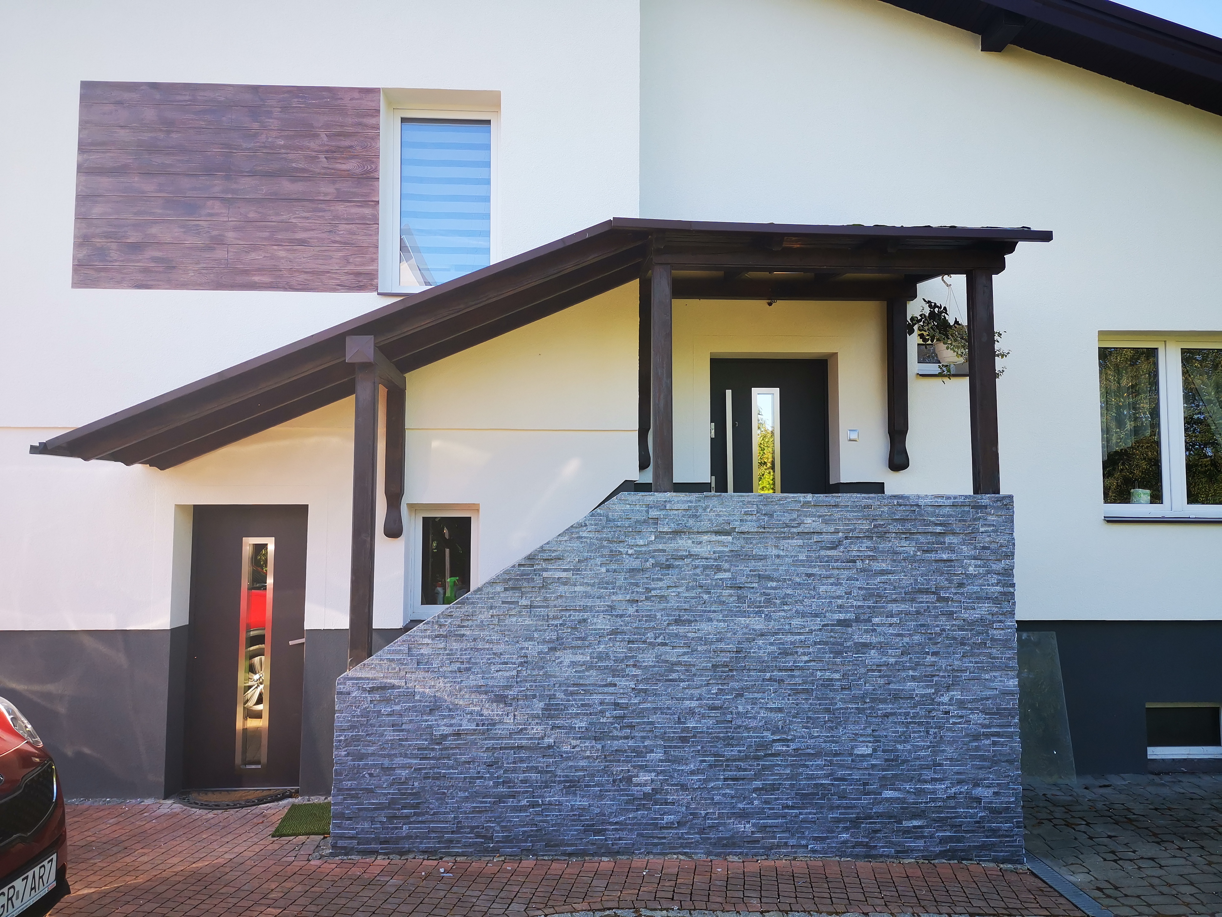Elewacja domu z kamienną okładziną schodów wejściowych i drewnianym panelem dekoracyjnym, widoczne okna z roletami i zadaszenie nad wejściem.