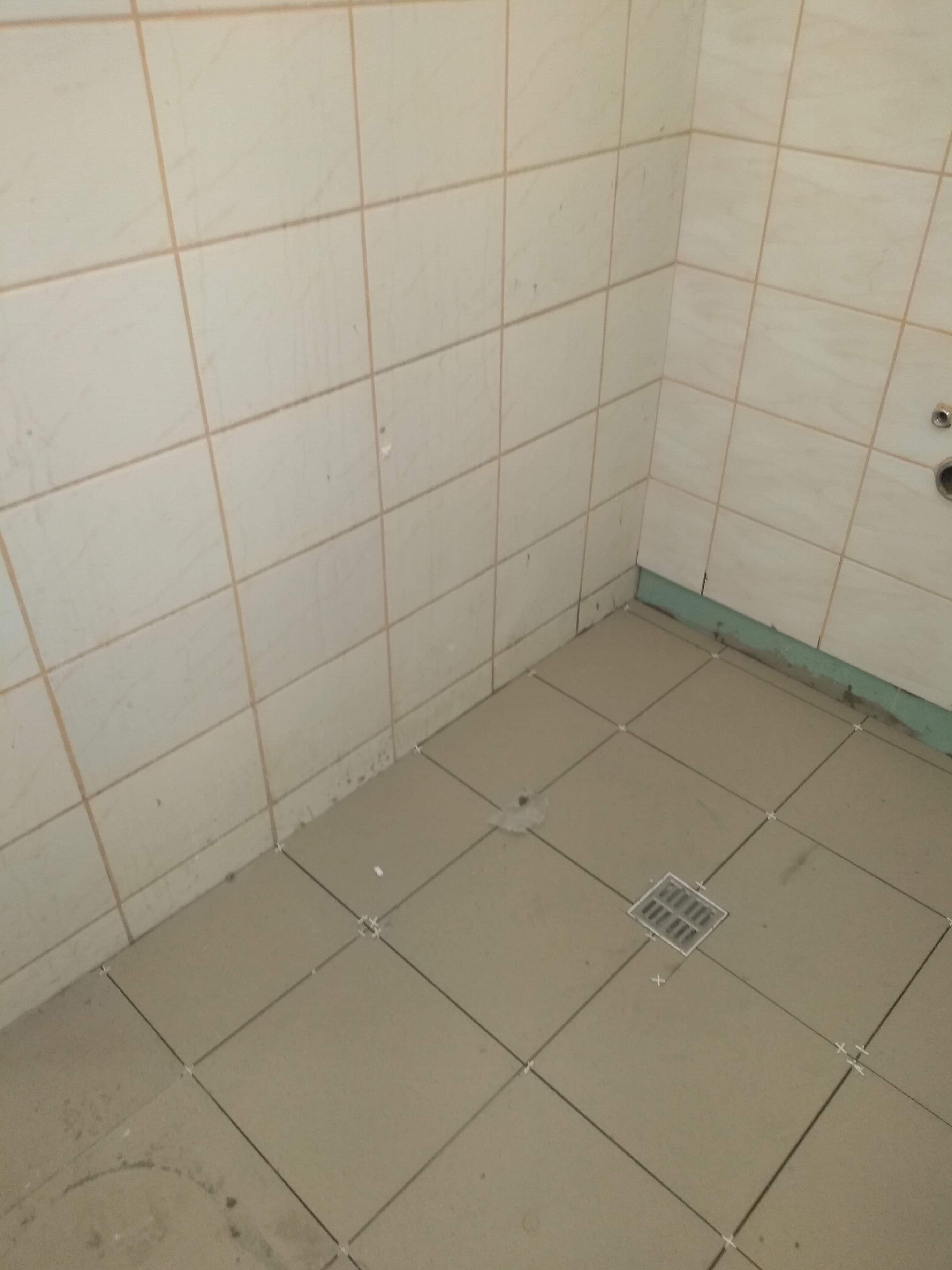 Fragment łazienki w trakcie układania płytek podłogowych i ściennych w beżowym odcieniu, widoczny odpływ liniowy i krzyżyki dystansowe.