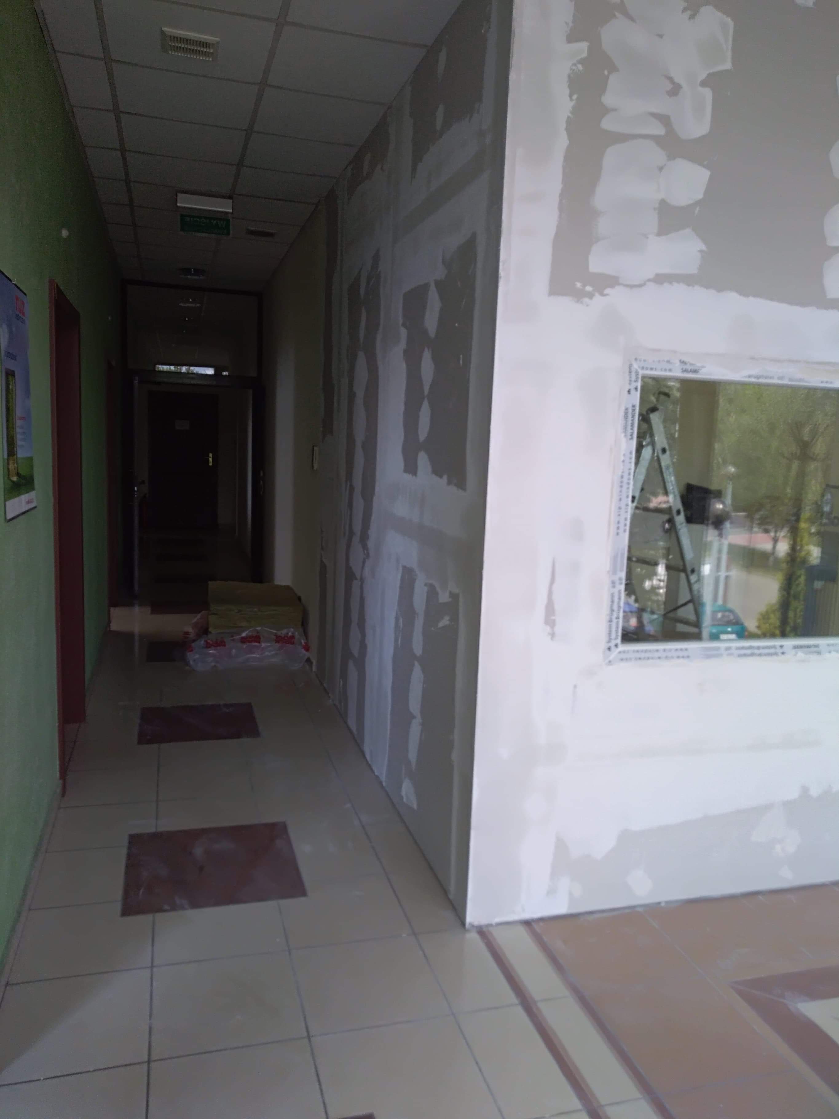 Korytarz w trakcie prac wykończeniowych, widoczne szpachlowane ściany, okno z drabiną opartą o parapet oraz fragment podłogi z płytek.
