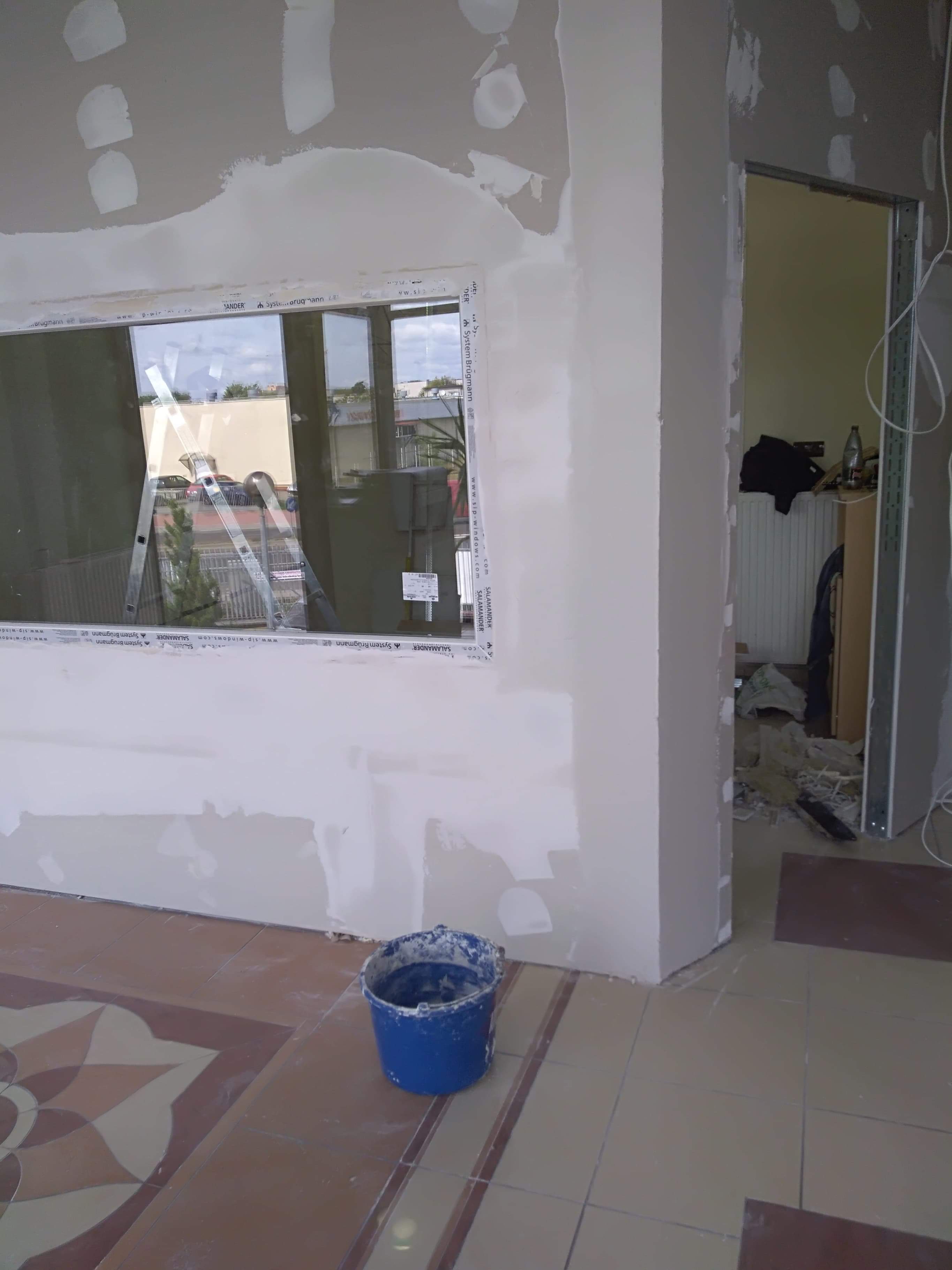 Ściana w trakcie szpachlowania z widocznymi łatami gładzi, niebieskie wiadro z zaprawą na ozdobnej, brązowo-beżowej podłodze z płytek, okno z odbijającą się drabiną w tle oraz widok na sąsiedni...