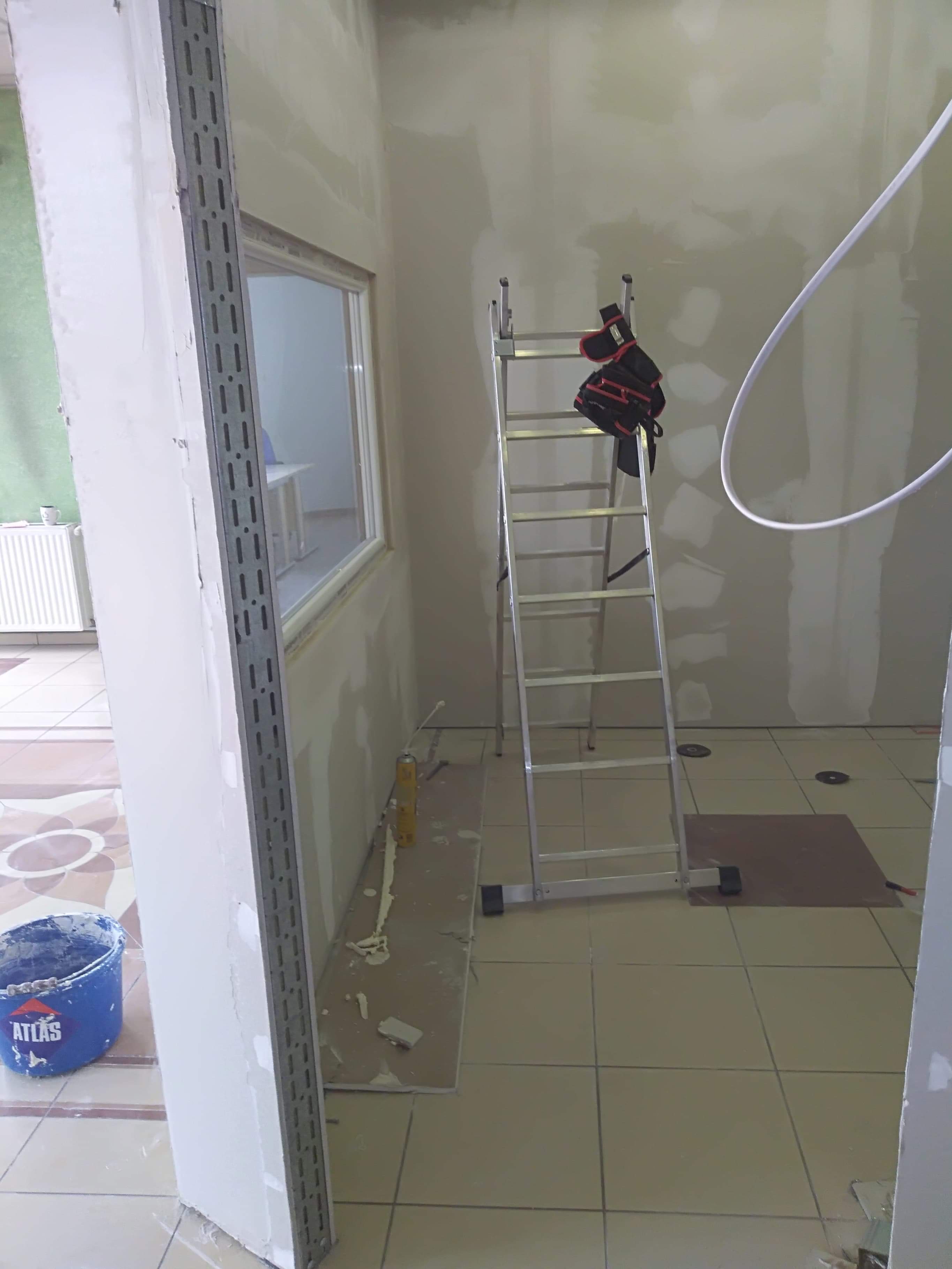 Wnętrze w trakcie prac przygotowawczych do malowania ścian, widoczna drabina z rękawicami roboczymi, wiadro z klejem Atlas, szpachlowane ściany i profile narożnikowe.