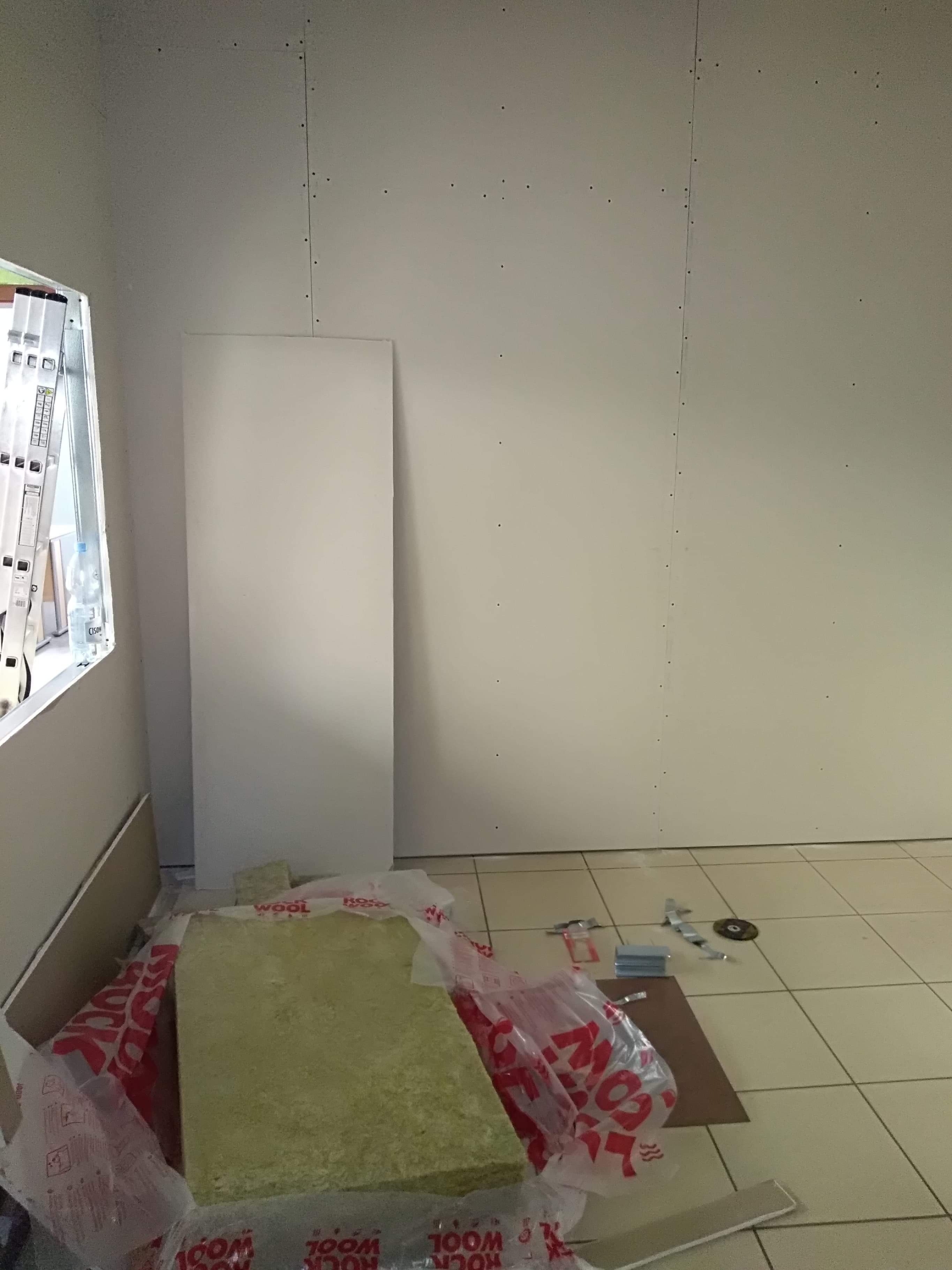 Wnętrze w trakcie montażu płyt gipsowo-kartonowych, widoczna wełna mineralna Rockwool w folii, drabina oparta o okno oraz narzędzia na podłodze.