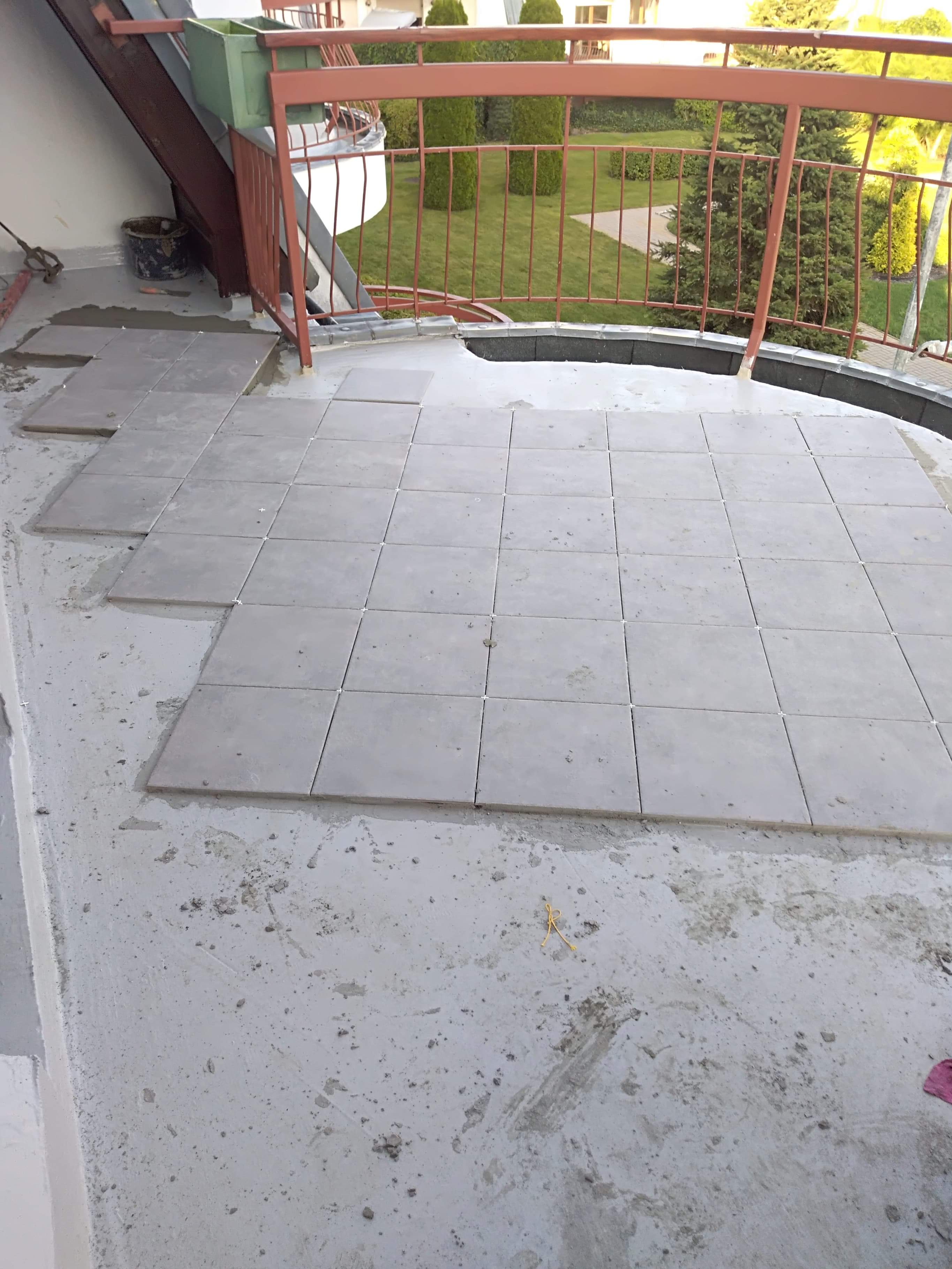 Układanie szarych płytek na balkonie z brązową balustradą, widok na ogród w tle, etap prac wykończeniowych.