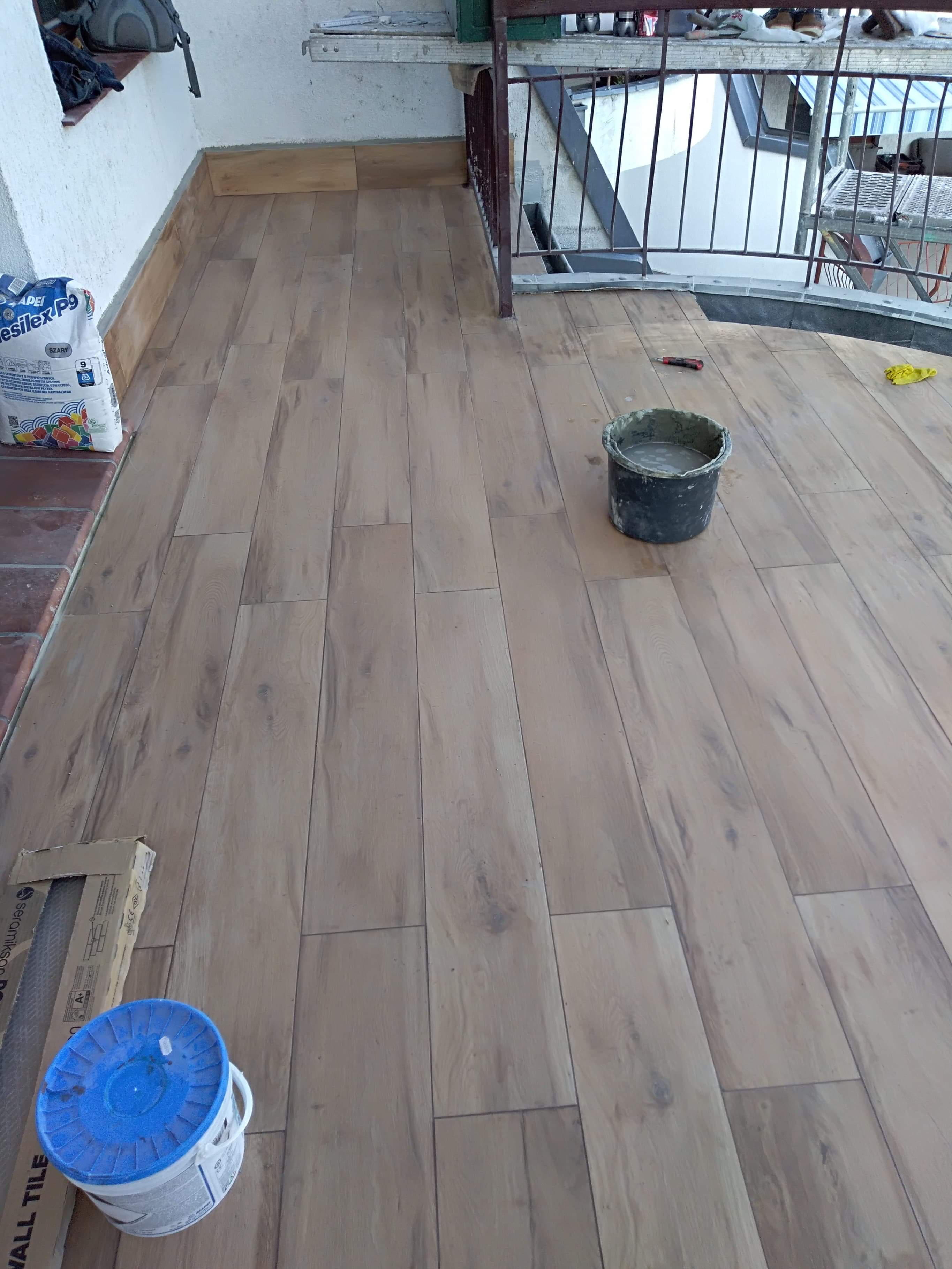 Balkon z nową, drewnopodobną podłogą z płytek, wiadro z zaprawą i worek kleju do płytek widoczne w trakcie prac.