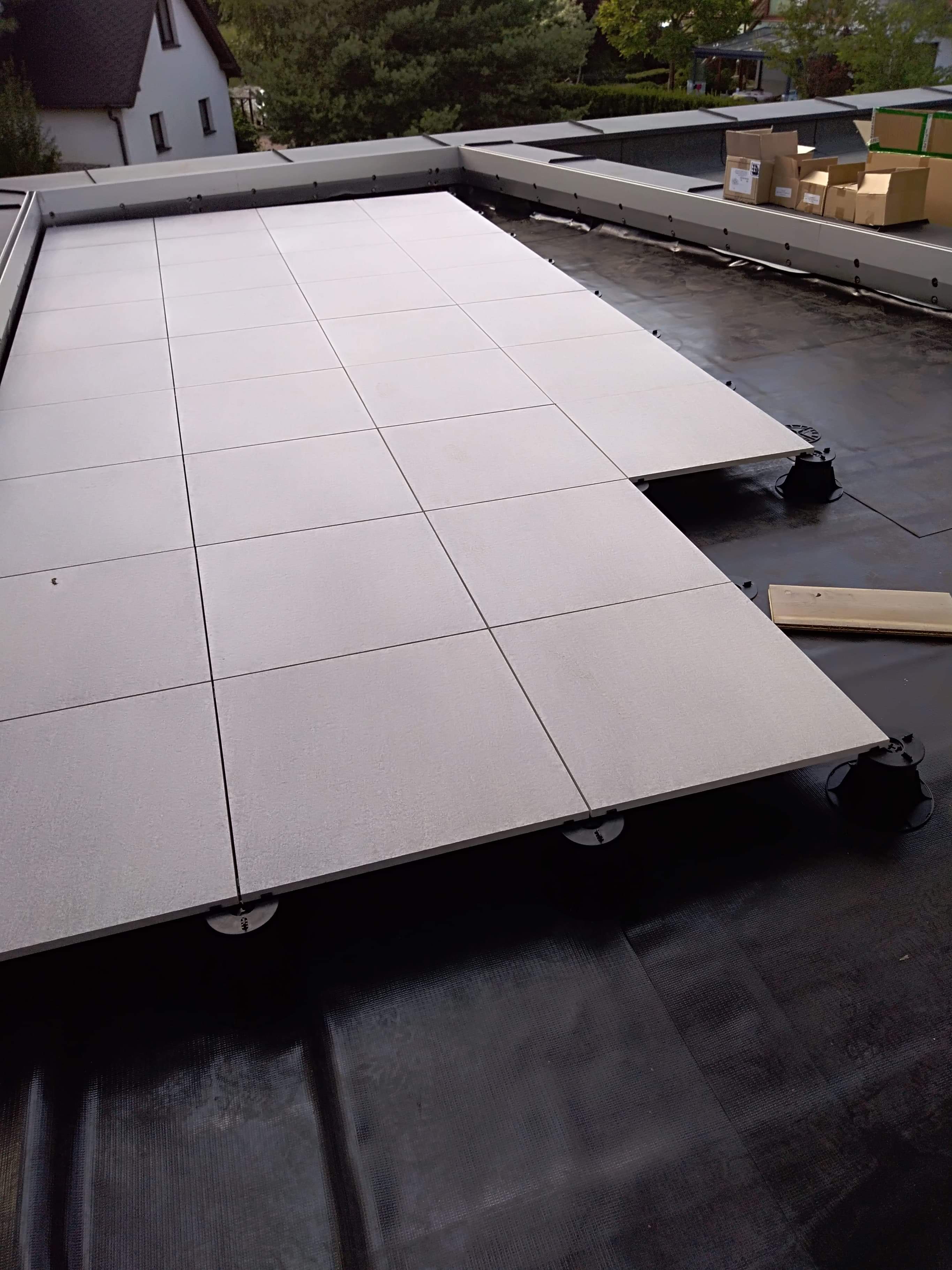 Montaż jasnoszarych płyt tarasowych na regulowanych wspornikach na czarnej hydroizolacji dachu płaskiego; w tle widoczne kartony i budynki.