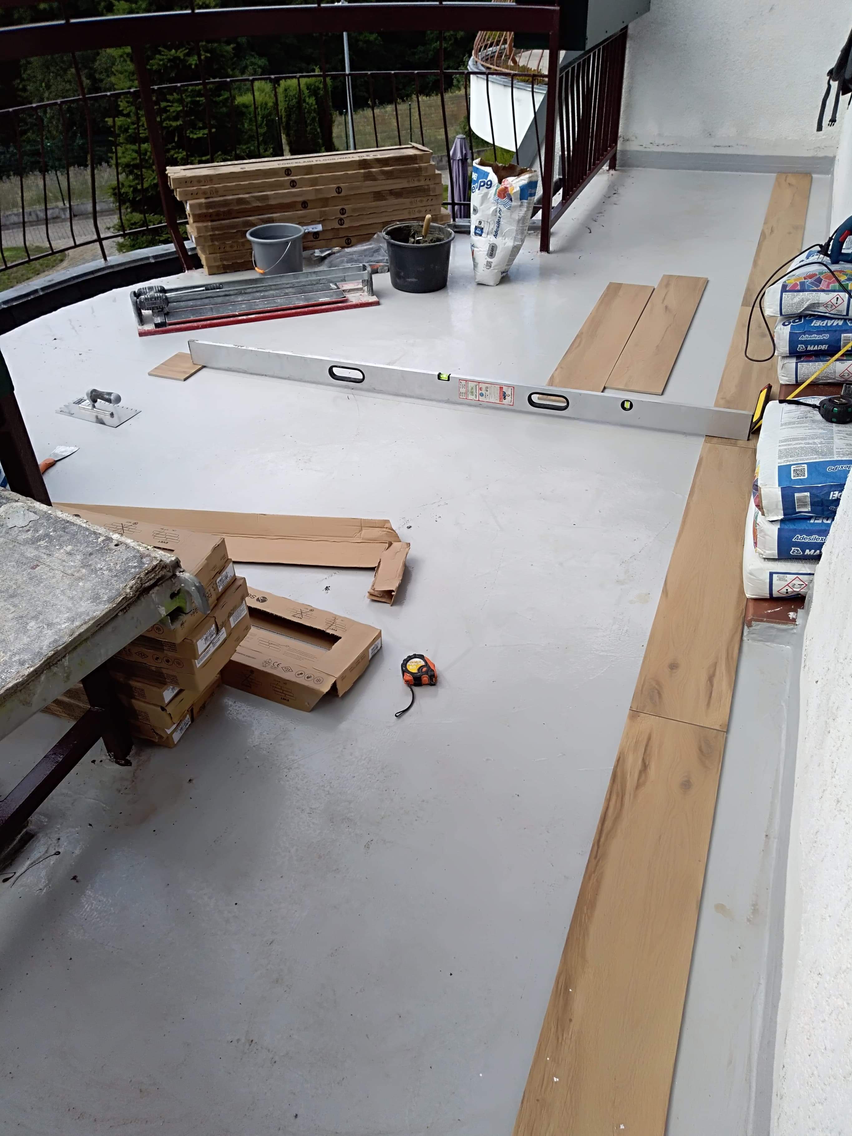 Przygotowanie do ułożenia drewnopodobnych płytek na balkonie: widoczne płytki, poziomica, wiadro z zaprawą, worki z klejem, przecinarka do płytek i miara.