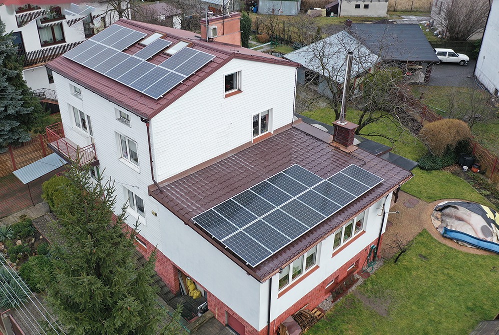 Dwukondygnacyjny dom z białymi ścianami i brązowym dachem, na którym zamontowano panele fotowoltaiczne, widok z lotu ptaka na posesję z ogrodem i budynkami gospodarczymi w tle.