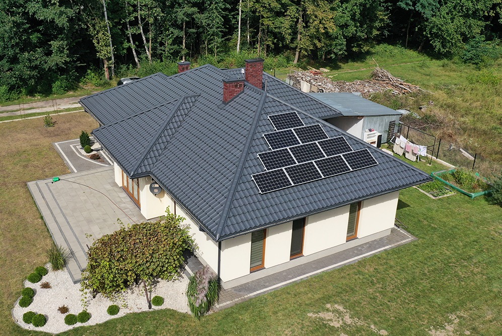Widok z góry na dom jednorodzinny z ciemnoszarą dachówką i panelami słonecznymi ułożonymi w dwóch rzędach na dachu, w tle widoczny ogród i sterta gałęzi.