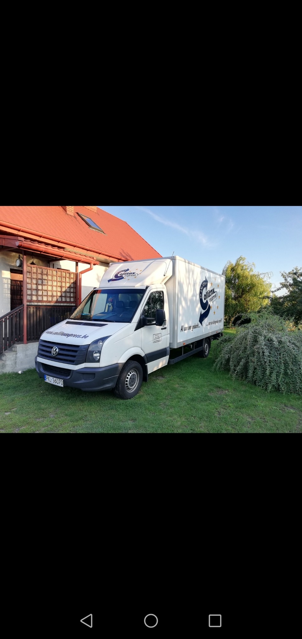 Biały bus kontenerowy marki Volkswagen z logo firmy transportowej 'Olitax Express' zaparkowany na trawniku przed domem z czerwonym dachem.