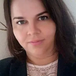 Portret kobiety o brązowych włosach i zielonych oczach, ubranej w koronkową bluzkę i czarną marynarkę, z delikatnym naszyjnikiem na szyi.