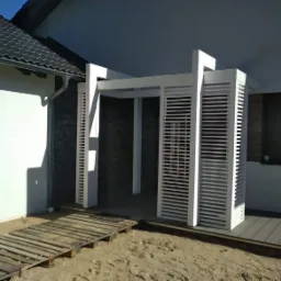 Nowoczesna, biała konstrukcja z lamelami, prawdopodobnie wejście na taras, z drewnianym podestem i prowizorycznym chodnikiem z palet, na tle domu z szarą dachówką.
