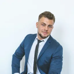 Młody mężczyzna w niebieskim garniturze, białej koszuli i czarnym krawacie, pozujący na jasnym tle, z widocznym zegarkiem na nadgarstku.