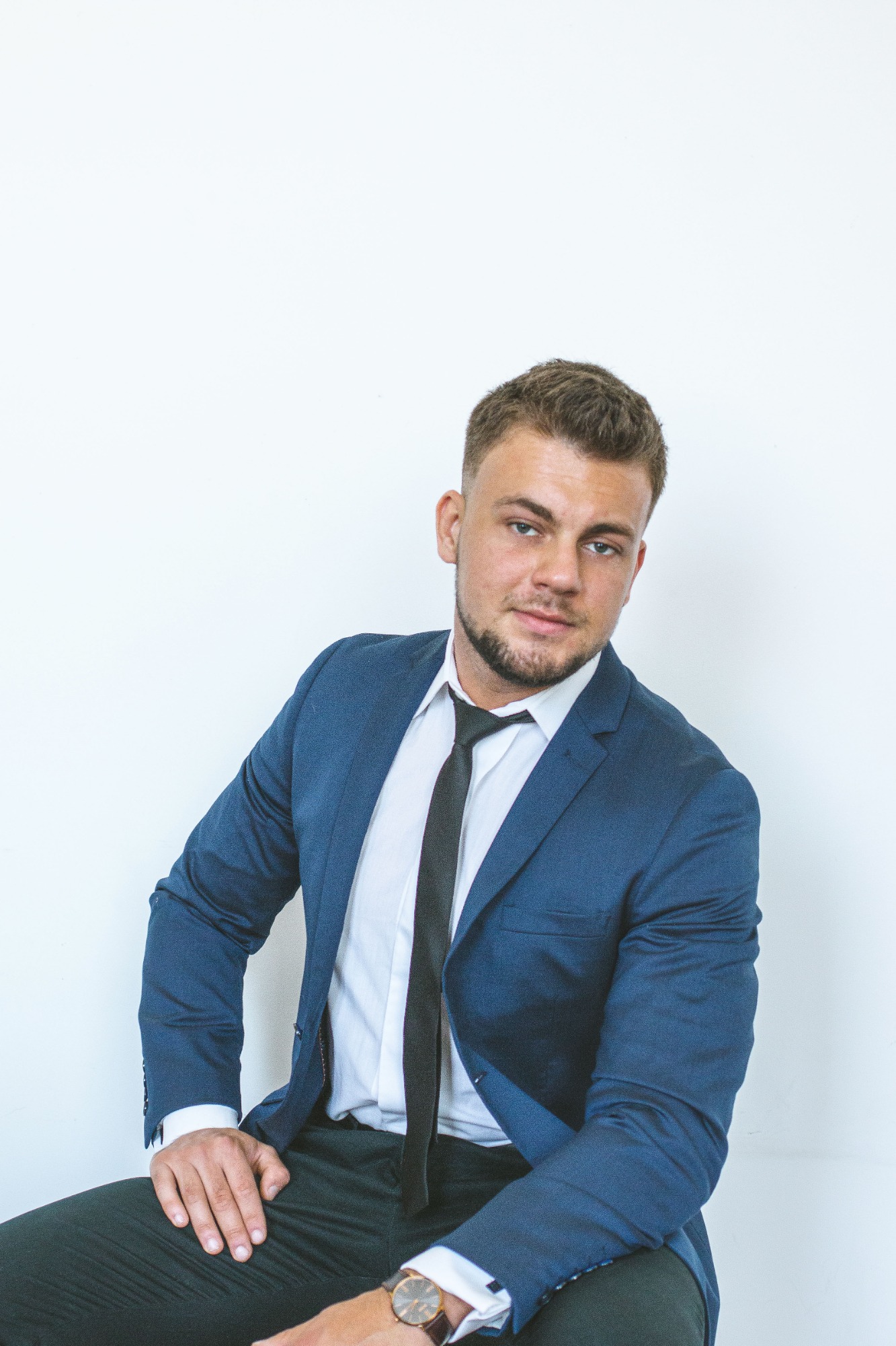 Młody mężczyzna w niebieskim garniturze, białej koszuli i czarnym krawacie, pozujący na jasnym tle, z widocznym zegarkiem na nadgarstku.
