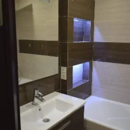 Nowoczesna łazienka z brązowo-beżowymi płytkami imitującymi drewno, podświetlane wnęki w ścianie, biała umywalka z szafką i chromowaną baterią, odbicie w lustrze fragmentu wanny.