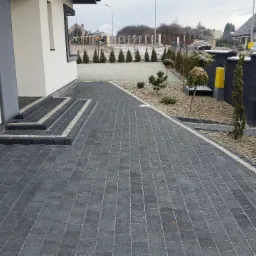 Ułożona ciemnoszara kostka brukowa przed nowoczesnym domem z trzema stopniami wejściowymi, obrzeża z jasnej kostki, w tle rząd tuj i szary płot z bramą.