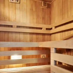 Wnętrze wykończonej drewnianej kabiny z ławkami, widoczny termometr, kratka wentylacyjna i oświetlenie pod ławkami.