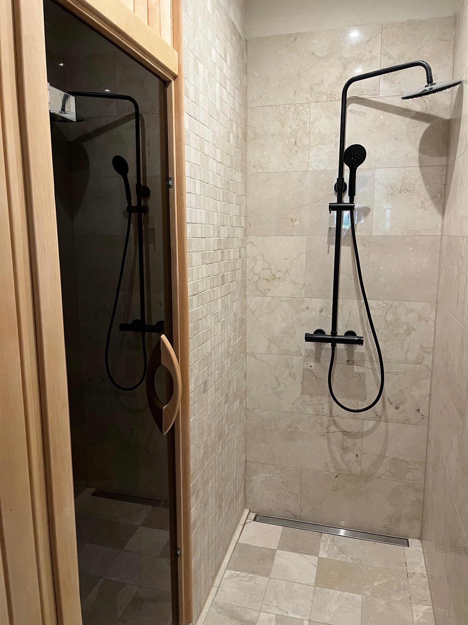 Wnętrze sauny z przeszklonymi drzwiami z drewna oraz czarnym zestawem prysznicowym na marmurowej ścianie.