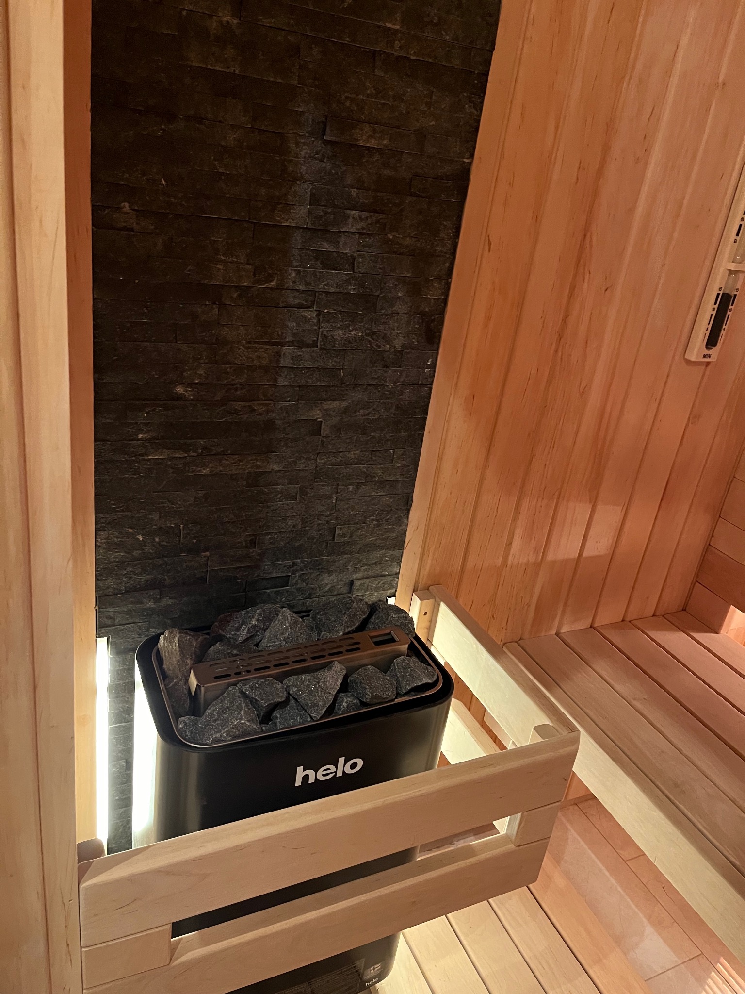 Wnętrze kabiny z drewnianymi ławkami, czarny piec z kamieniami marki Helo podświetlony od dołu, ściana z ciemnego kamienia.
