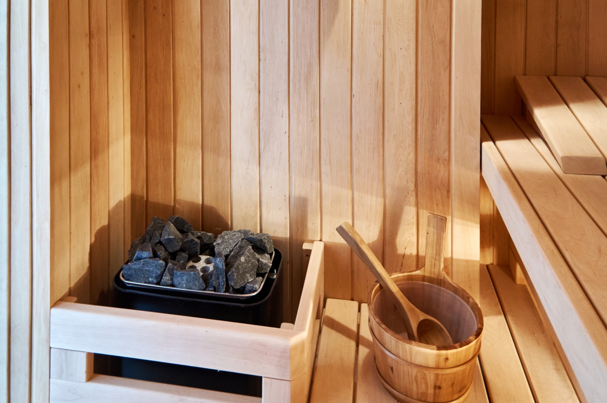 Wnętrze wykończonej drewnem sauny z kamieniami w piecu i drewnianym czerpakiem w wiadrze.