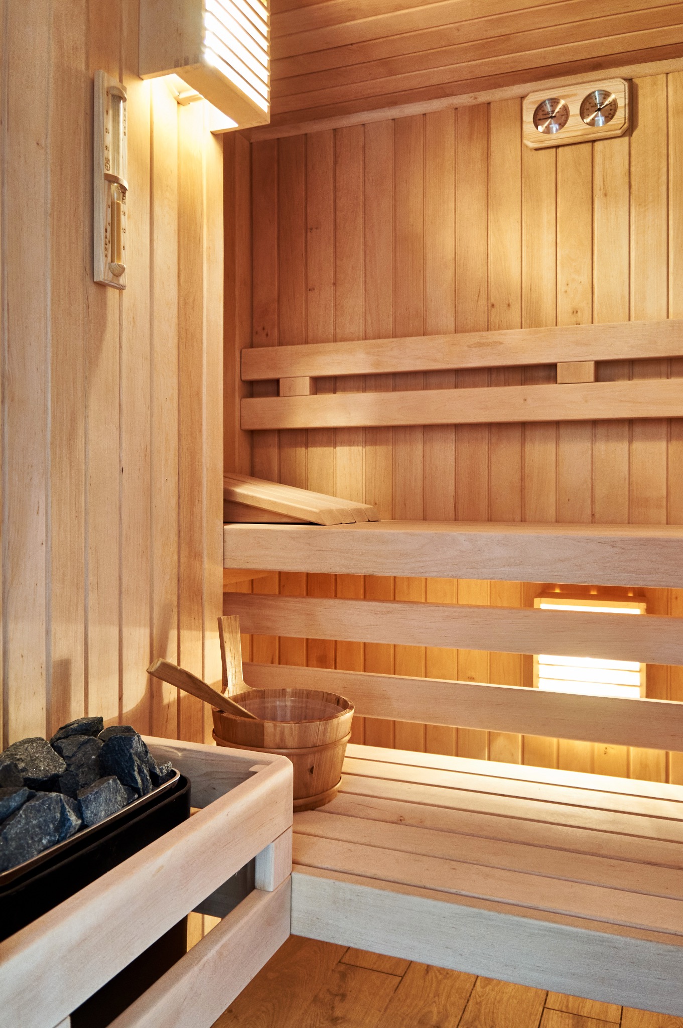 Wnętrze jasnej, drewnianej sauny w Warszawie z kamieniami w piecu, drewnianym wiadrem z chochlą i termometrem ściennym.