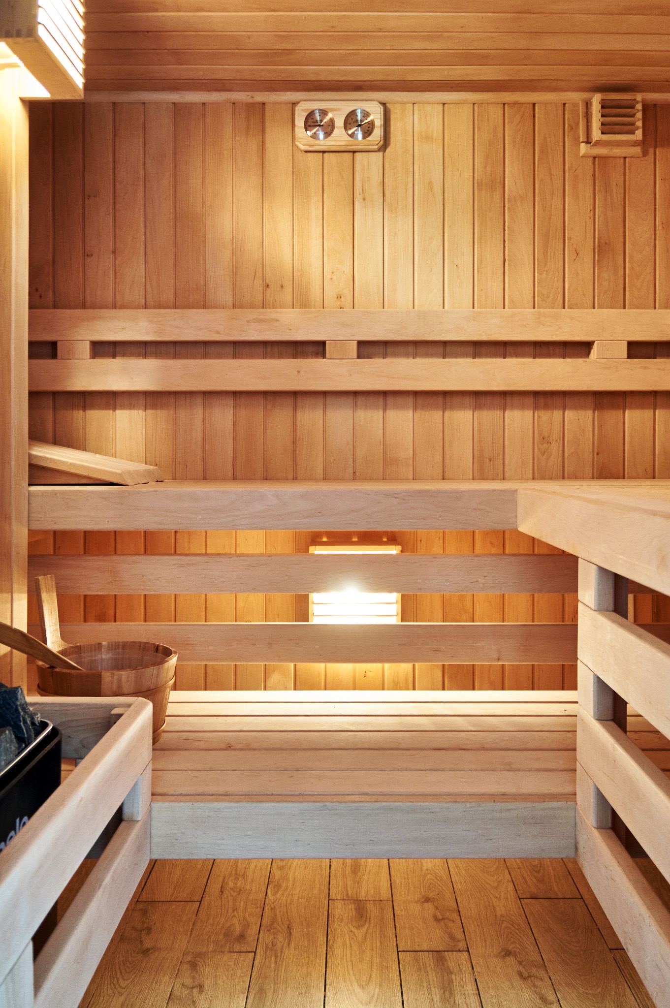 Wnętrze drewnianej kabiny z ławkami, wiadrem i kamieniami w koszyku, oświetlone ciepłym światłem, z widocznymi termometrami na ścianie.