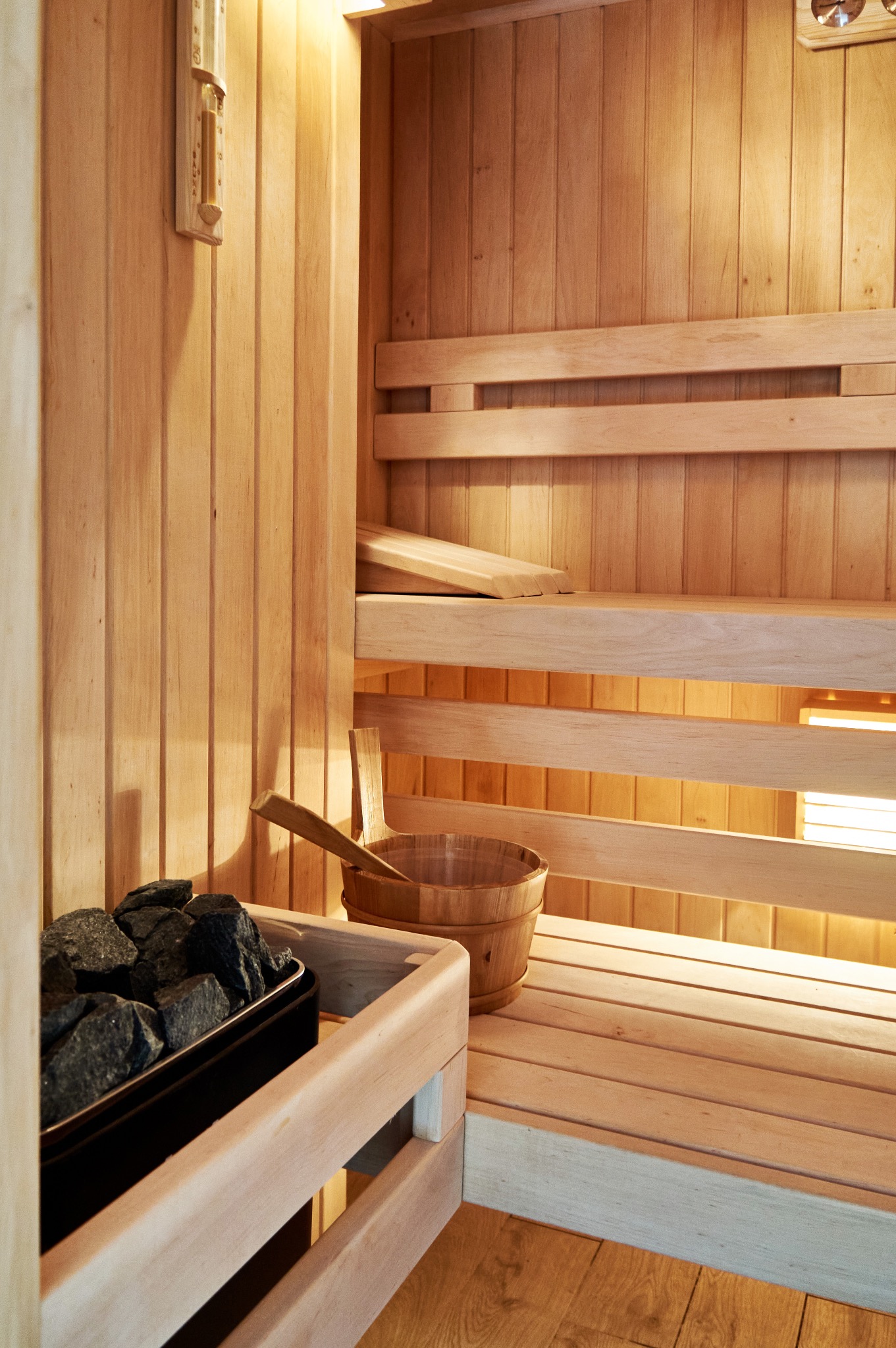 Wnętrze sauny z jasnego drewna z kamieniami w pojemniku, drewnianym wiadrem i termometrem na ścianie.