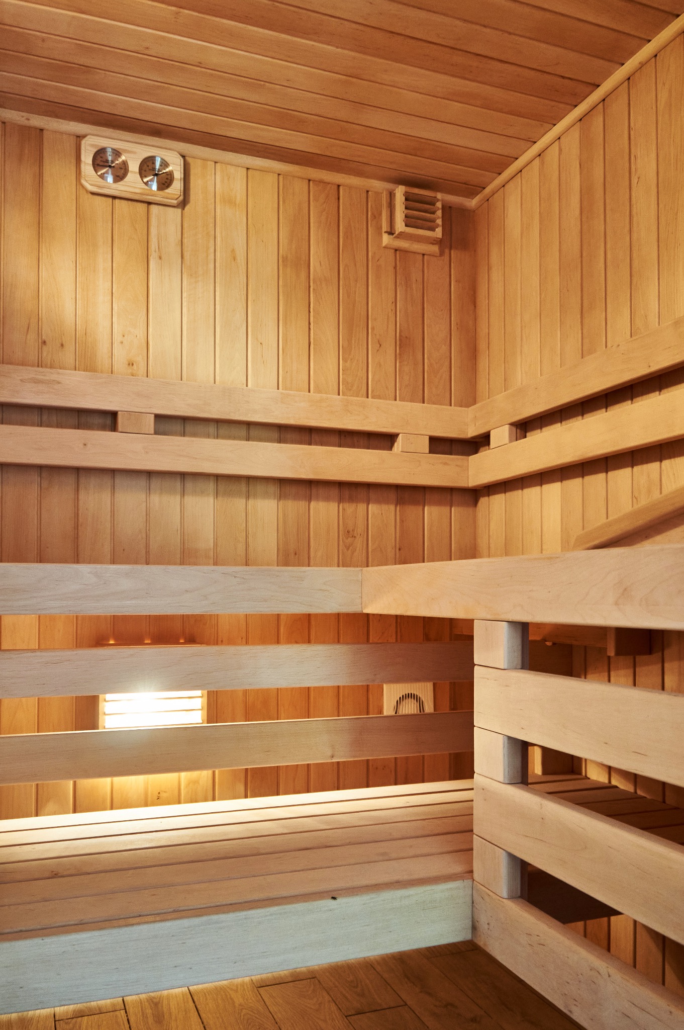 Wnętrze wykończonej drewnianej kabiny z ławkami, widoczny termometr, kratka wentylacyjna i oświetlenie pod ławkami.