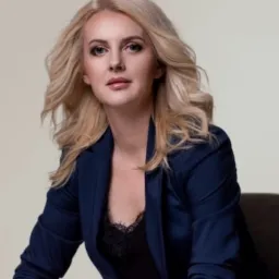 Portret kobiety w granatowym żakiecie, z blond włosami i delikatnym makijażem, siedzącej na ciemnym fotelu na neutralnym tle.