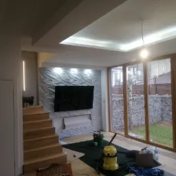 Wnętrze pokoju po remoncie, z panelami 3D na ścianie za telewizorem, drewnianymi schodami, dużymi oknami z widokiem na ogród i sprzętem do sprzątania po remoncie.