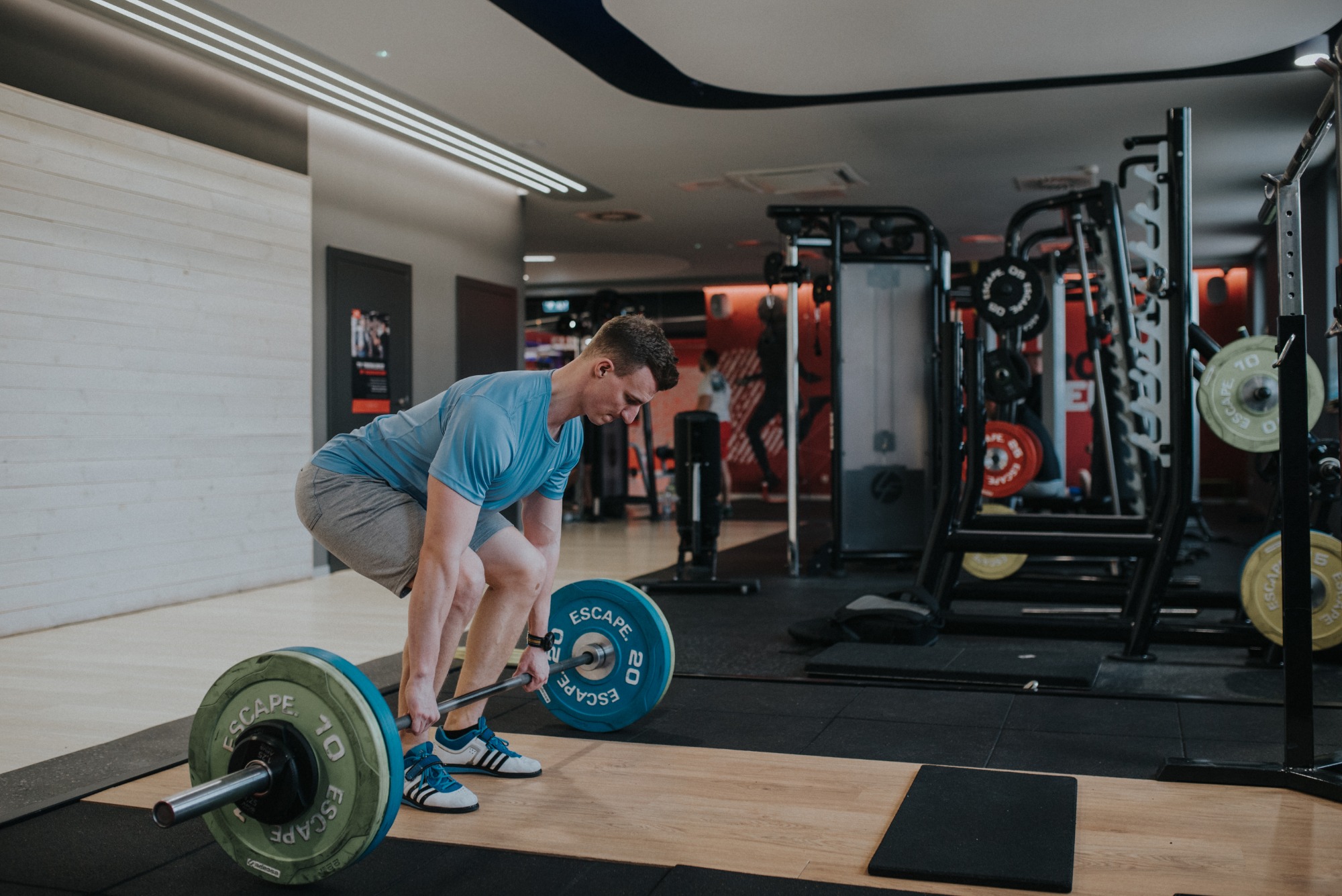 Mężczyzna w sportowym stroju wykonuje martwy ciąg w siłowni, używając sztangi z kolorowymi obciążeniami Escape Fitness.
