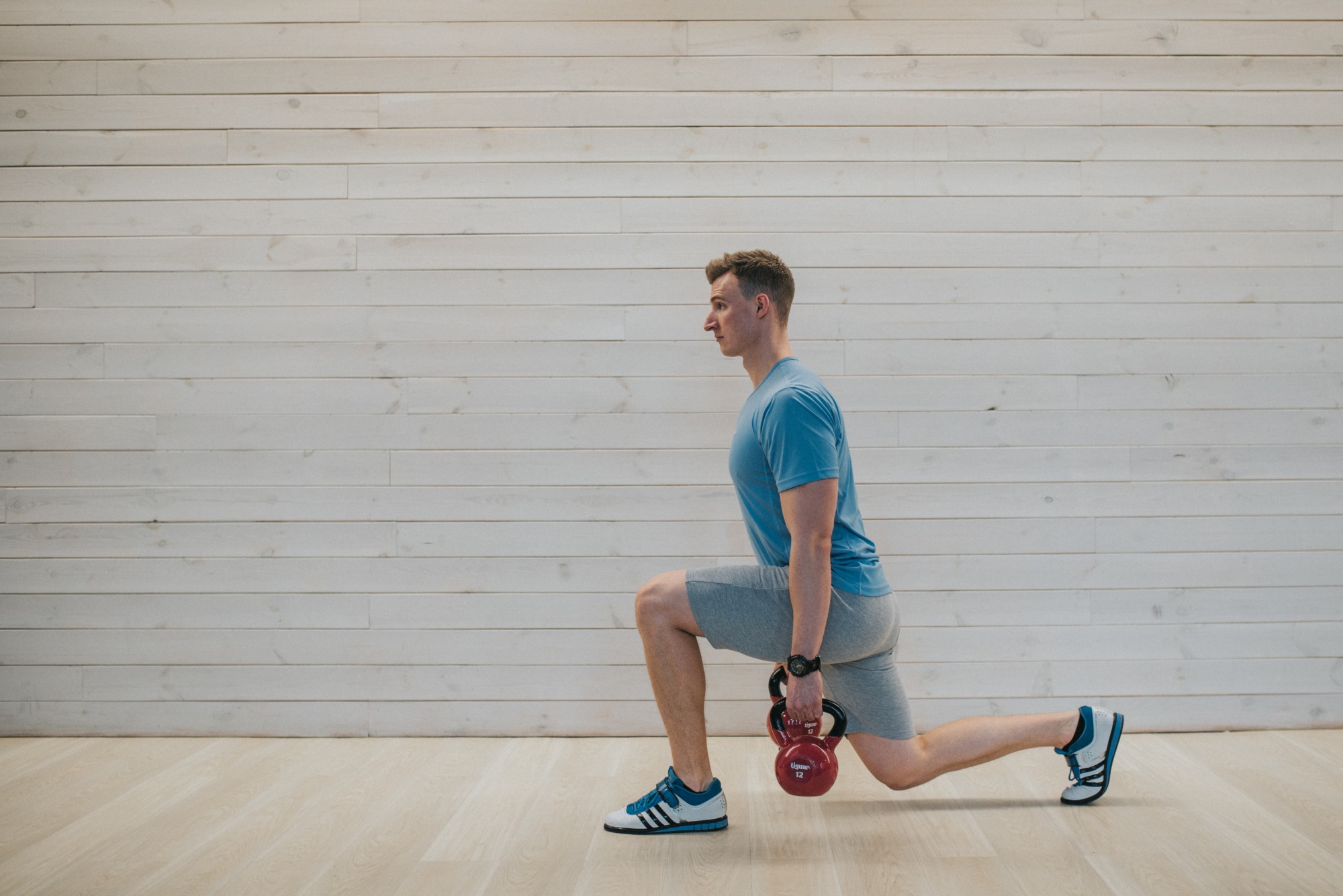 Mężczyzna w niebieskiej koszulce i szarych spodenkach wykonuje wypad z czerwonym kettlebellem w dłoniach, na tle białej drewnianej ściany.