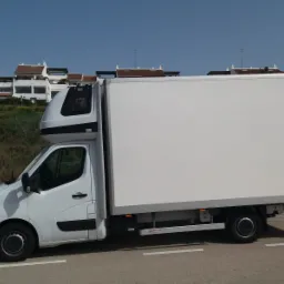 Biały samochód dostawczy z nadbudówką kabiny na tle zabudowy mieszkalnej i zieleni, widziany z boku, zaparkowany na ulicy.