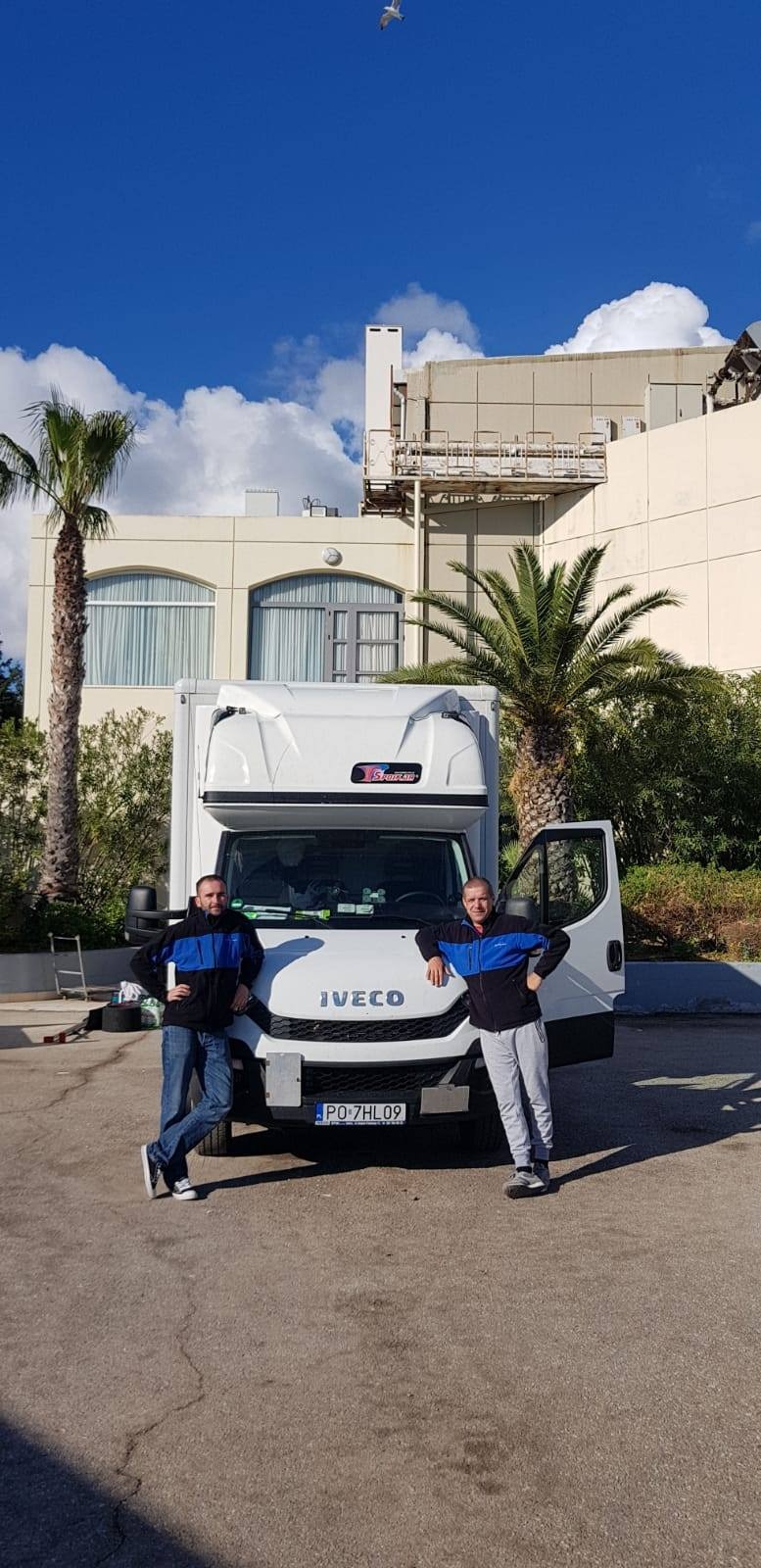 Dwóch mężczyzn w kurtkach pozuje na tle białej furgonetki Iveco z otwartymi drzwiami w słoneczny dzień, z palmami i budynkiem w tle oraz ptakiem na niebie.