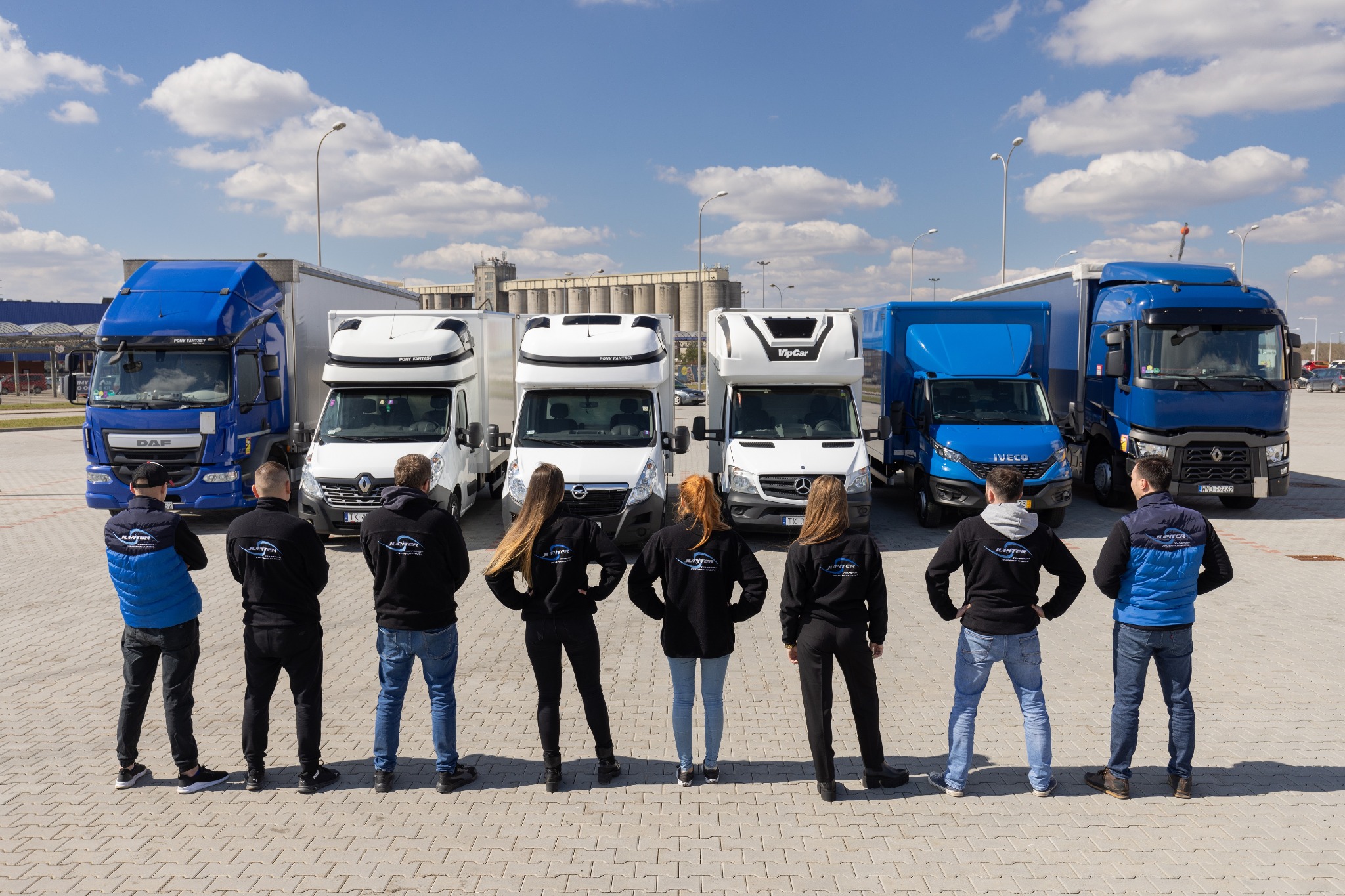 Flota pojazdów firmy transportowej Jupiter, składająca się z pięciu samochodów dostawczych i ciężarowych w kolorach białym i niebieskim, ustawionych w rzędzie na placu z kostki, z pracownikami...